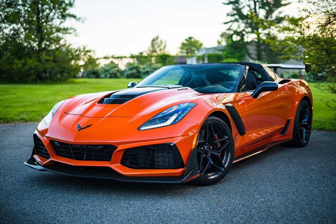 Bright orange luxury sports car parked on a driveway with trees and grass in the background.